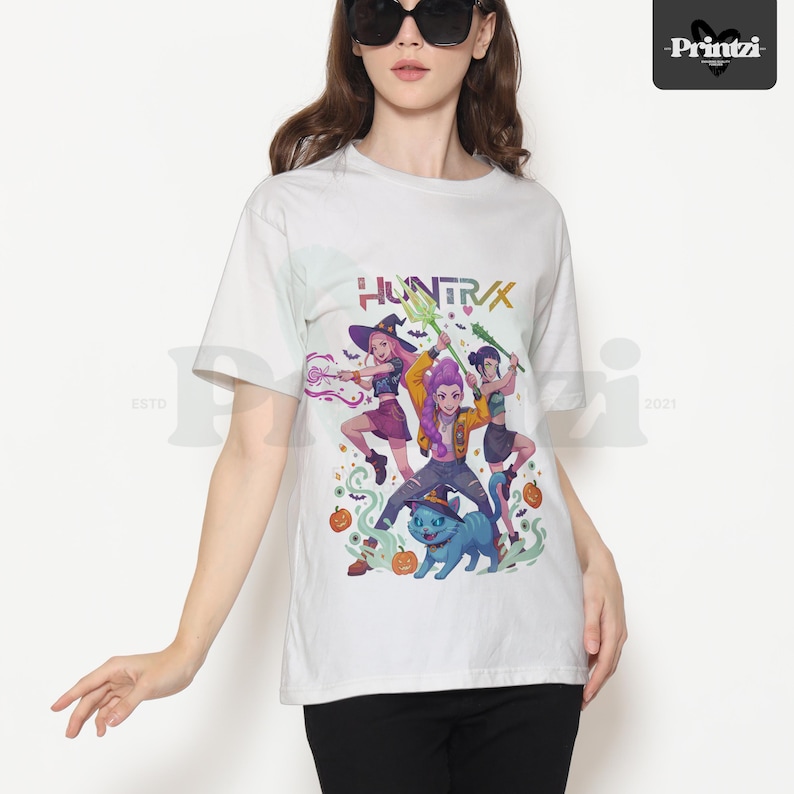 May include: White t-shirt featuring a colorful graphic of three animated characters with weapons, a blue cat, and pumpkins. The word "HUNTRX" is printed above the characters. The shirt is a crew neck style.