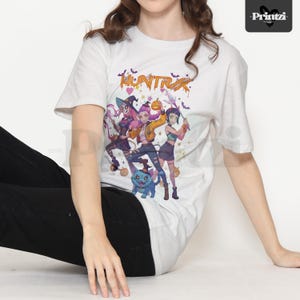 May include: White t-shirt featuring a colorful graphic of three anime-style characters in Halloween costumes, with the word "HUNTRX" above them. The design includes bats, pumpkins, and a blue cat. The shirt is worn by a person.