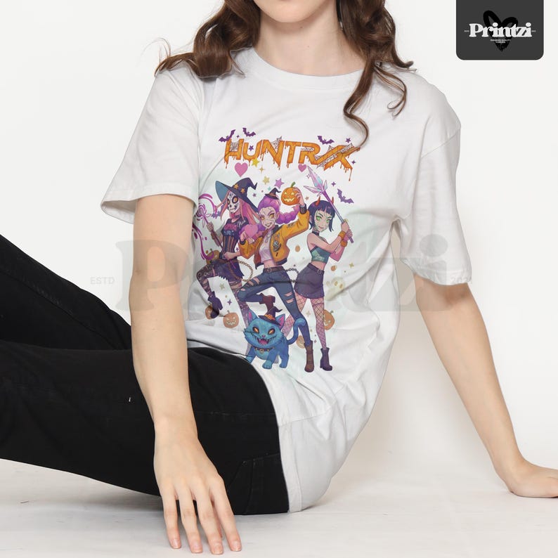 May include: White t-shirt featuring a colorful graphic of three anime-style characters in Halloween costumes, with the word "HUNTRX" above them. The design includes bats, pumpkins, and a blue cat. The shirt is worn by a person.