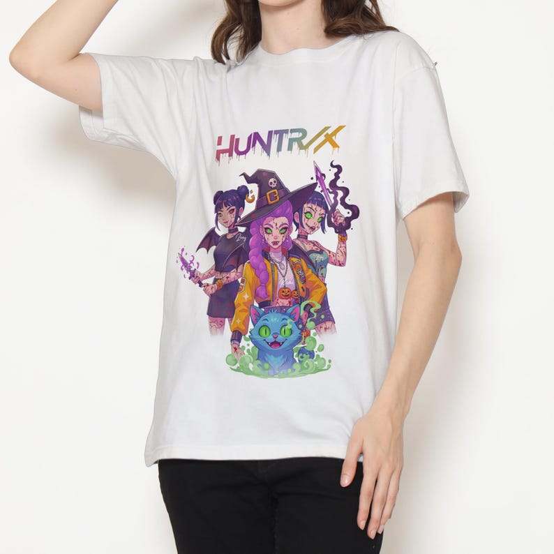 May include: White t-shirt featuring a colorful graphic of three stylized witches and a blue cat. The word "HUNTRIX" is printed above the graphic in a rainbow gradient. The witches are holding various magical items.