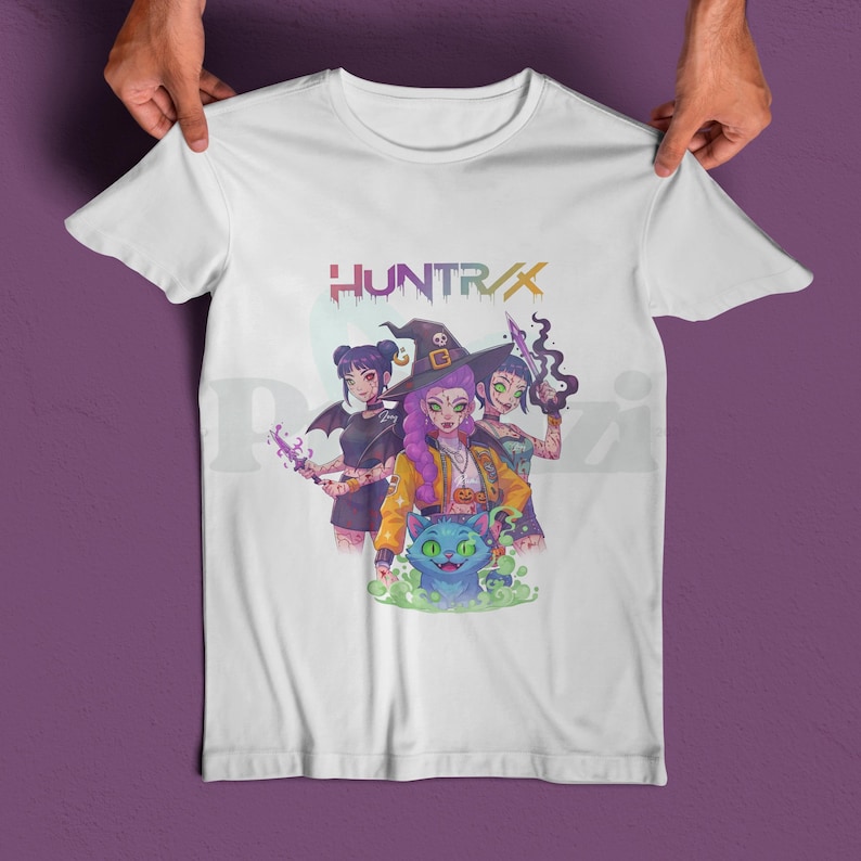 May include: White t-shirt featuring a colorful graphic of three witches with a cat and the text "HUNTR/X". The witches are in a cartoon style, with purple, yellow, and green accents. The cat is blue and green.
