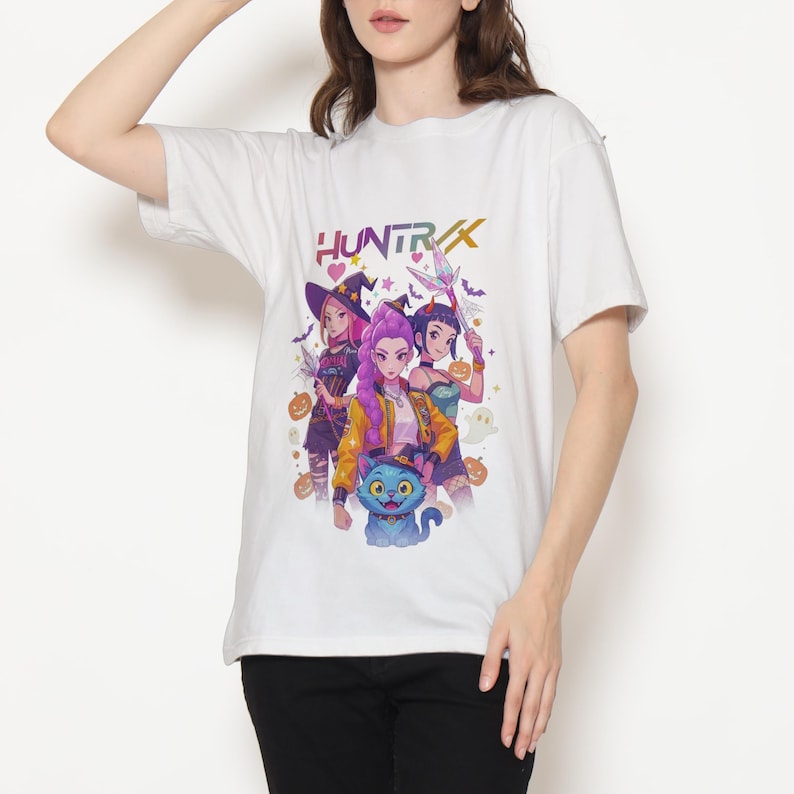 May include: White t-shirt featuring a colorful graphic of three anime-style characters with a blue cat and the word "HUNTRX". The design includes pumpkins, stars, and witch-themed elements. The shirt is made of a soft, comfortable material.