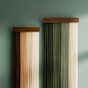 May include: Two wall hangings with a wooden top and cascading strands. One features a gradient of brown to beige, the other green to beige. They are mounted on a teal wall, adding a touch of modern decor to the space.