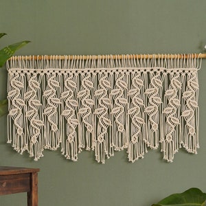 May include: A white macrame wall hanging with a geometric pattern, hanging from a wooden dowel rod against a sage green wall.