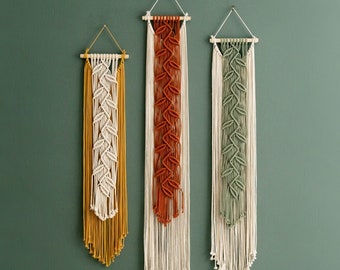 May include: Three macrame wall hangings with different colored patterns. The hangings are made of natural fibers and have a bohemian style. The hangings are hung on a green wall.