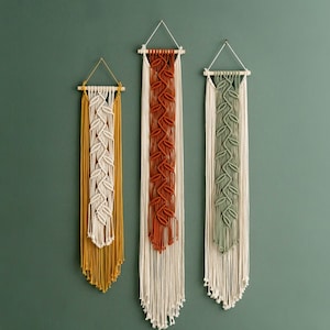 May include: Three macrame wall hangings with different coloured patterns. The hangings are made of natural fibres and have a bohemian style. The hangings are hung on a green wall.