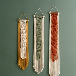 Puede incluir: Tres tapices de macramé de pared con diferentes patrones de colores. El tapiz de la izquierda es amarillo, el del medio es verde y el de la derecha es naranja.