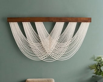 May include: A macrame wall hanging featuring a dark brown wooden bar with numerous off-white cords arranged in a semi-circular pattern. The wall behind the art is painted a muted green color. The piece is a decorative home accent.