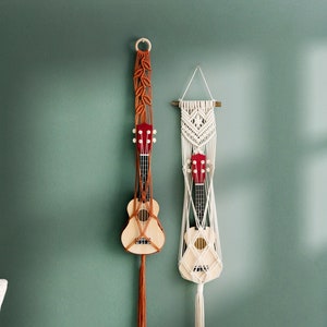 May include: Two macrame wall hangings with wooden dowels, each holding a ukulele. One hanging is brown with a leaf pattern and the other is white with a geometric pattern.