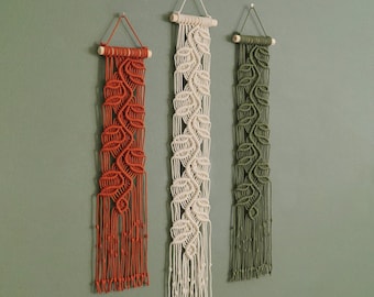May include: Three vertical macrame wall hangings suspended from wooden dowels. Each piece features a central vine-like pattern with leaf-shaped knots. The left hanging is rust orange, the center is cream, and the right hanging is olive green.