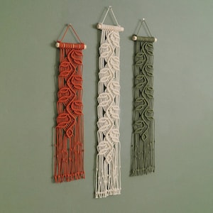 Puede incluir: Tres tapices de macramé de pared con un patrón de hojas. Los tapices son en tonos naranja, blanco y verde.