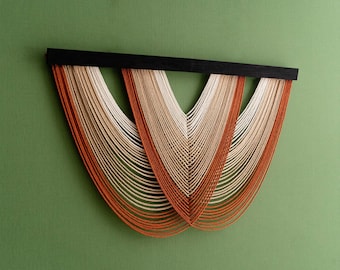 May include: A contemporary wall hanging with a black wooden bar and flowing strands in white, beige, and orange hues. The strands are arranged in a layered, curved pattern against a green backdrop, forming a unique, artistic piece for home decor.