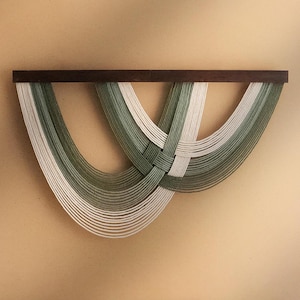 May include: A wall hanging featuring a dark brown wooden shelf with woven strands of white and green thread. The threads are arranged in a symmetrical pattern, creating a decorative and textured design. The wall art is mounted on a light tan wall.