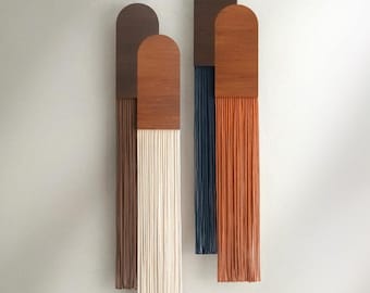 May include: Four vertical wall hangings with wooden tops and fringed bottoms. The tops are arched and made of dark brown and natural wood. The fringe colours are brown, cream, navy blue, and orange. They are mounted on a light grey wall.