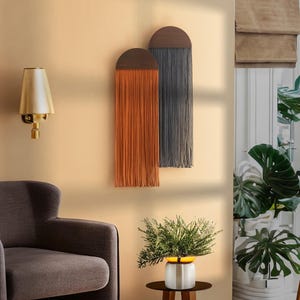 May include: Two wall hangings made of yarn with a half-circle wooden top. The left hanging is orange and the right hanging is gray. The hangings are on a beige wall with a lamp and a plant in the foreground.