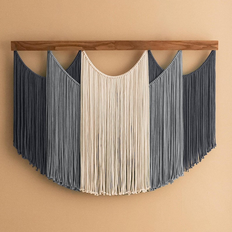 May include: A macrame wall hanging with a wooden bar. The design features three sections of hanging cords in shades of dark gray, light gray, and cream. The cords are arranged in a curved, symmetrical pattern.