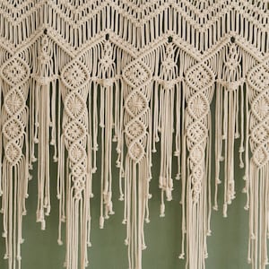 May include: A white macrame wall hanging with a geometric pattern and fringe. The hanging is made of cotton cord and has a bohemian style.