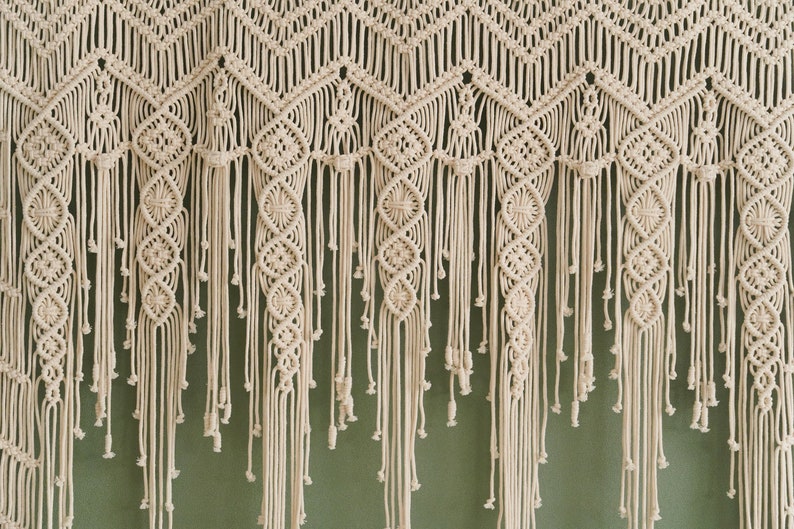 May include: A white macrame wall hanging with a geometric pattern and fringe. The hanging is made of cotton cord and has a bohemian style.