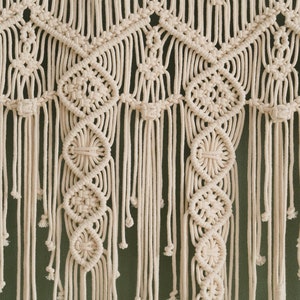 May include: A white macrame wall hanging with a geometric pattern. The hanging is made of cotton cord and has a fringe at the bottom.