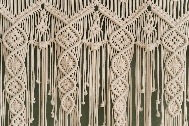 May include: A white macrame wall hanging with a geometric pattern. The hanging is made of cotton cord and has a fringe at the bottom.