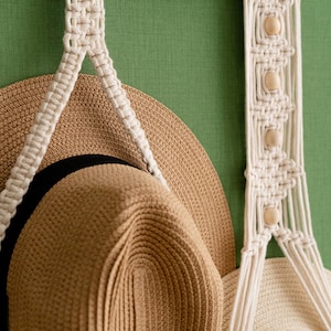 May include: A macrame hat rack with a natural wood dowel and off-white cotton cord. The rack features a woven design with wooden beads and holds two straw hats with black and white bands. The background is a solid green.