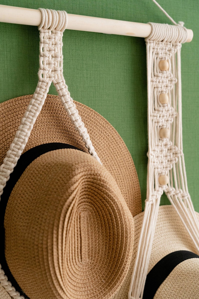 May include: A macrame hat rack with a natural wood dowel and off-white cotton cord. The rack features a woven design with wooden beads and holds two straw hats with black and white bands. The background is a solid green.