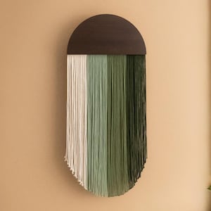May include: A decorative wall hanging featuring a dark brown, semi-circular wooden top and vertical yarn strands in white, light green, medium green, and dark green. The yarn strands create a gradient effect, adding texture to the piece.