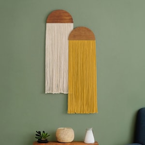 May include: Two macrame wall hangings with wooden tops. The left hanging is white and the right hanging is yellow. The hangings are on a green wall with a wooden shelf below.