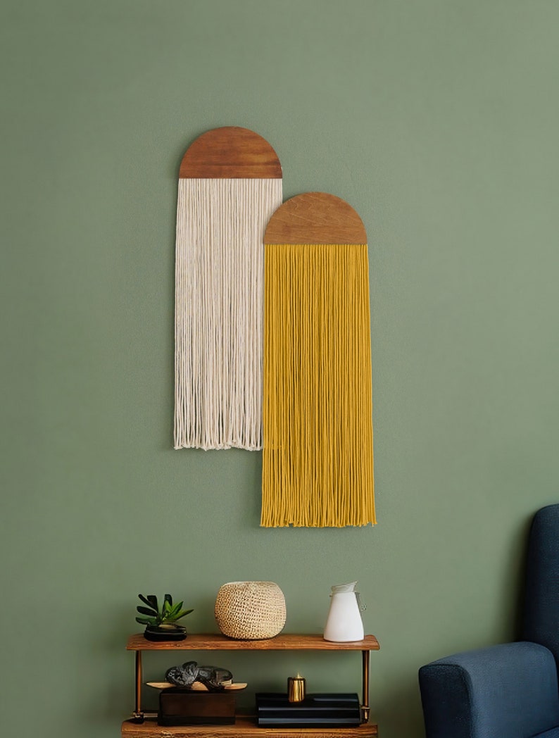 May include: Two macrame wall hangings with wooden tops. The left hanging is white and the right hanging is yellow. The hangings are on a green wall with a wooden shelf below.