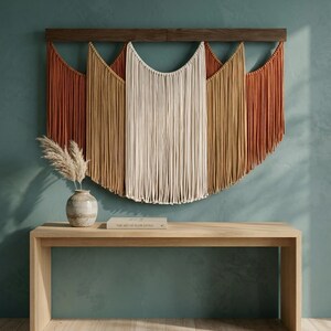 May include: A large macrame wall hanging with a wooden bar, featuring a color palette of rust, tan, and cream. Below, a light wood console table with a vase and a book titled "The Art of Slow Living".