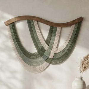 May include: A wall hanging featuring a curved, dark wood support bar from which multiple strands of sage green and cream-coloured cord are suspended. The cords are arranged in overlapping, U-shaped layers, creating a rhythmic, layered pattern.
