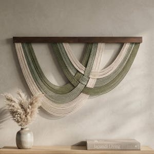 May include: A wall hanging featuring multiple draped strands of sage green and cream-colored rope suspended from a dark brown horizontal wooden bar. The ropes are arranged in overlapping, curved, U-shaped patterns that create a layered effect.