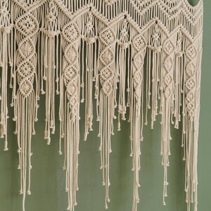 May include: A white macrame wall hanging with a geometric pattern and fringe. The hanging is made of cotton cord and is attached to a wooden dowel.