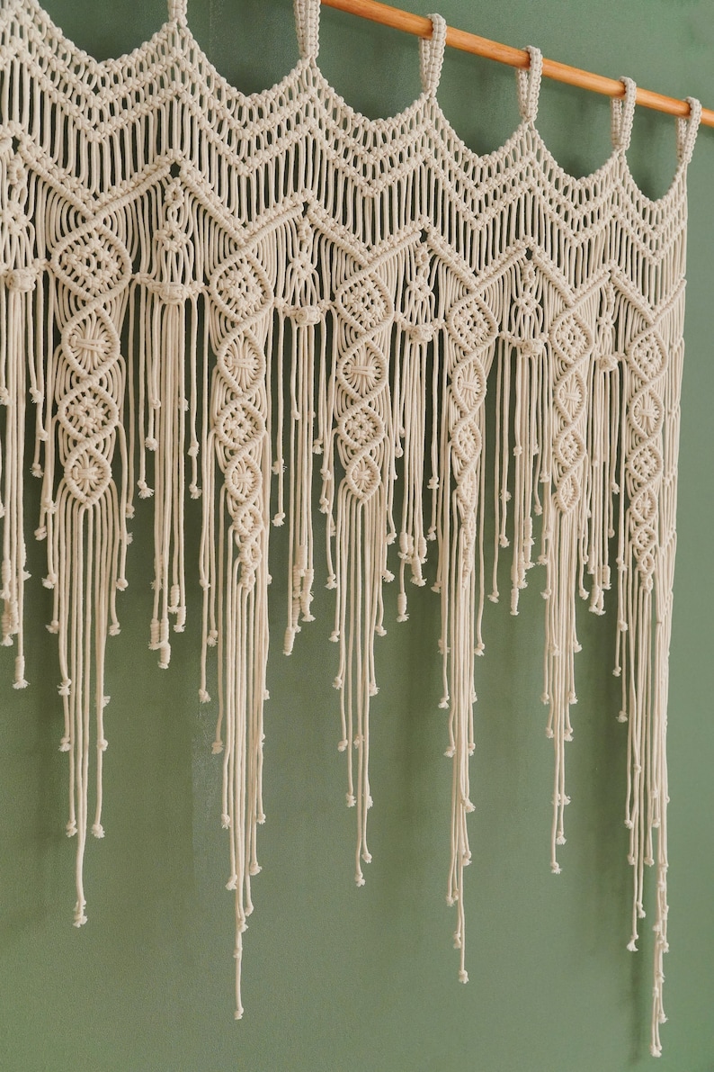 May include: A white macrame wall hanging with a geometric pattern and fringe. The hanging is made of cotton cord and is attached to a wooden dowel.