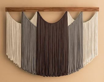May include: A macrame wall hanging with a wooden rod. The design features a gradient of colors, transitioning from off-white to gray and ending with a deep brown. The fringe hangs in a curved, symmetrical pattern, creating a modern, textured wall decor.