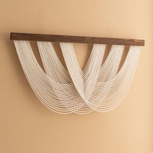 以下が含まれることがあります： 濃い茶色の木の棒から、多数のオフホワイトのコードが垂れ下がっているモダンな壁掛け。 コードは対称的で流れるようなパターンに配置され、テクスチャのある芸術的なデザインを作り出しています。 壁掛けはベージュの壁に取り付けられています。
