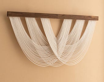 May include: A modern wall hanging featuring a dark brown wooden bar from which numerous strands of off-white cord cascade. The cords are arranged in a symmetrical, flowing pattern, creating a textured, artistic design. The wall hanging is mounted on a beige wall.