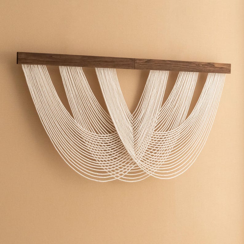 May include: A modern wall hanging featuring a dark brown wooden bar from which numerous strands of off-white cord cascade. The cords are arranged in a symmetrical, flowing pattern, creating a textured, artistic design. The wall hanging is mounted on a beige wall.