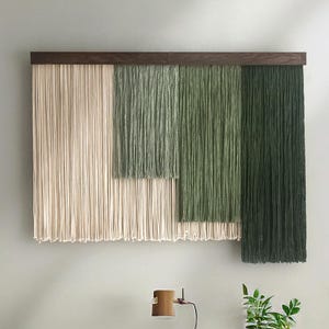 May include: A contemporary wall hanging with a dark wood frame and a textured design. The artwork displays vertical strands in cream, light green, medium green, and dark green, forming a layered, geometric pattern. This piece adds a touch of natural texture to any room.