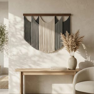 May include: A large, neutral-toned macrame wall hanging with a dark wood top. The design features a series of vertical fringe in shades of gray and cream. The wall hanging is mounted above a light wood console table. A vase with dried flowers sits on the table.