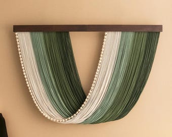 May include: A wall hanging featuring a dark wooden bar with vertical strands of green and white yarn. The yarn is arranged in a gradient pattern, with a row of small, white pom-poms along the curved bottom edge. The wall hanging is mounted on a beige wall.