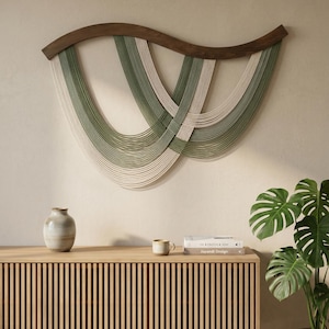 May include: A modern wall hanging featuring a curved, dark wood top with woven strands in shades of green and off-white. The artwork hangs above a light wood cabinet with vertical slats, a vase, and a plant.