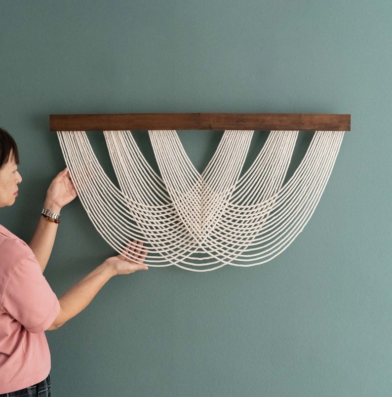 May include: A macrame wall hanging with a dark brown wooden bar and off-white cords. The cords are arranged in a curved, symmetrical pattern, creating a decorative, textured design. The wall hanging is displayed against a teal-colored wall.