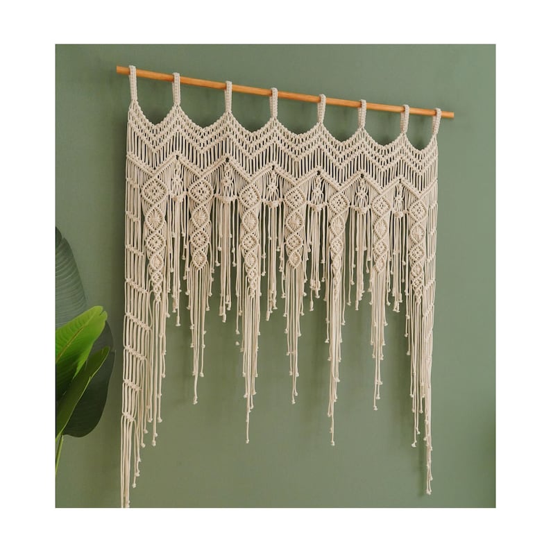 May include: A white macrame wall hanging with a geometric pattern and fringe. The hanging is attached to a wooden dowel rod.