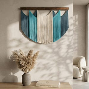 May include: A large, semi-circular wall hanging with a wooden top and cascading fringe in shades of blue and cream. The wall decor is mounted on a light-colored wall, creating a focal point in a modern living space.