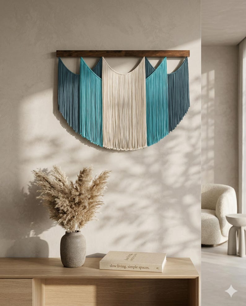 May include: A large, semi-circular wall hanging with a wooden top and cascading fringe in shades of blue and cream. The wall decor is mounted on a light-colored wall, creating a focal point in a modern living space.