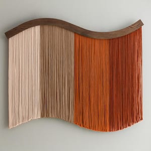 May include: A decorative wall hanging featuring a curved wooden top and vertical strands in beige, brown, and orange. The strands create a flowing, wave-like pattern. This textured piece adds warmth and visual interest to a room.