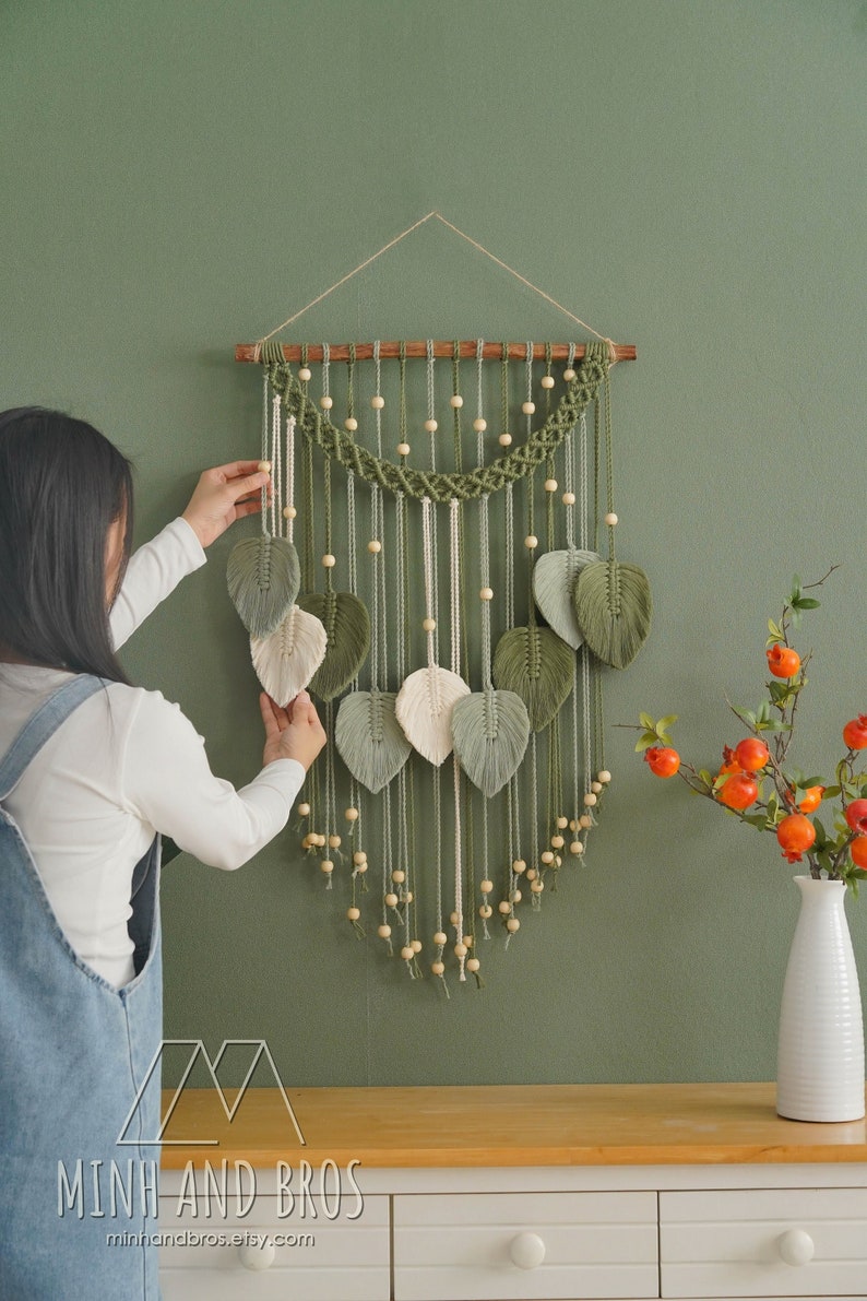 May include: A green and white macrame wall hanging with wooden beads and leaf shapes. The hanging is on a green wall and a person is adjusting the leaves.