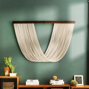 May include: A white macrame wall hanging with a wooden dowel rod, hanging on a sage green wall. The macrame is in the shape of a V, with the strings crossing in the middle.