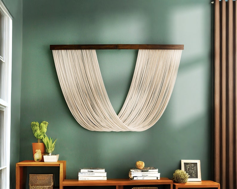 May include: A white macrame wall hanging with a wooden dowel rod, hanging on a sage green wall. The macrame is in the shape of a V, with the strings crossing in the middle.